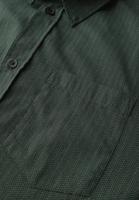 Camisa de botones de color verde oscuro con un patrón de espiga, confeccionada en una tela suave, con un bolsillo en el pecho y botones negros.