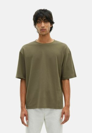 Junger Mann mit dunklen Haaren, der ein olivgrünes, übergroßes T-Shirt mit kurzen Ärmeln und helle Hosen trägt, im Stehen vor einem schlichten Hintergrund.