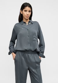 Η γκρι μακρυμάνικη μπλούζα διαθέτει κούμπωμα με κουμπιά και τσέπη, είναι κατασκευασμένη από λεία ύφασμα και παρουσιάζει ανάγλυφο μοτίβο με το λογότυπο σε όλη την επιφάνεια.