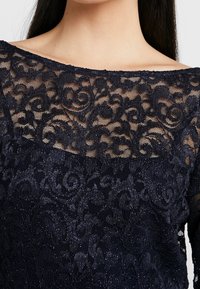 Námořnická krajkov á blůza se květinovým vzorem, lodičkovým výstřihem a průhlednou texturou s jemným leskem, zdůrazňujícími složité detaily.