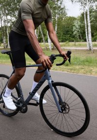 Vélo de canyon gris avec roues noires, freins à disque et guidon en baisse. Le cycliste porte un t-shirt vert, un short noir et des chaussures blanches.