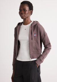 Brun zip-up hettegenser laget av mykt materiale, med en brystlomme med logo, kombinert med en hvit ribbestrikket skjorte og svarte bukser.