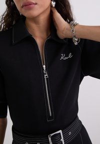 Top zippé noir à manches courtes, avec des coutures blanches contrastées et "Karl" brodé. Stylé avec une ceinture large et des boucles d'oreilles décoratives.