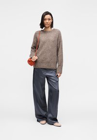 Καφέ πλεκτό πουλόβερ, άνετη εφαρμογή, σε συνδυασμό με γυαλιστερό σκούρο γκρι παντελόνι με φαρδιά μπατζάκια. Το μοντέλο κρατά μια μικρή πορτοκαλί τσάντα. Απλό, ουδέτερο φόντο.