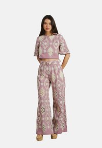 Gekürztes pinkes Oberteil mit kunstvollen weißen Mustern, kombiniert mit weiten pinken Hosen, die ähnliche Designs und ein gebundenes Taillendetail aufweisen.