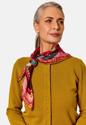 Zijde sjaal met gedurfde rode, roze en groene patronen, stijlvol om de nek gedrapeerd over een mosterdkleurige cardigan met knopen.