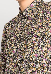 Blouse florale avec un fond noir, ornée d'un mélange de fleurs jaunes, roses et vertes. Design col avec des accents de boutons.