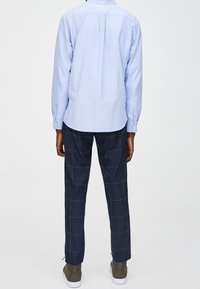 Hellblaues, langärmliges Hemd mit Knöpfen, Rückansicht; kombiniert mit karierten, dunkelblauen Hosen mit grünen Linien. Legerer, entspannter Schnitt.