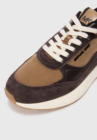 Sneaker featuring a brown suede and textured fabric upper, with a white lace closure, and a beige outsole. Branding located on the tongue.