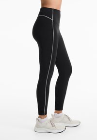 Black leggings with a high waistband, featuring a white contrasting seam along the side. Paired with light-colored athletic shoes.