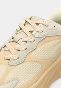Beige sneakers med mesh-paneler og læderdetaljer. Har lyse orange snørebånd og trykt branding på siden. Struktureret overflade overalt.