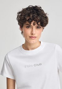 Vit bomull t-shirt med rund halsringning, korta ärmar och en silverfärgad "Polo Club"-logotyp tryckt på framsidan. Slät textur.