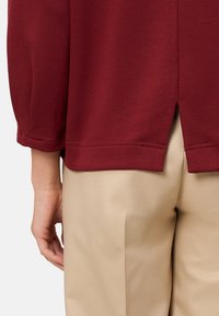 Bordo, długą bluzkę z rozciętym dołem, w połączeniu z beżowymi spodniami. Materiał jest gładki, z widocznym przeszyciem wzdłuż brzegów.