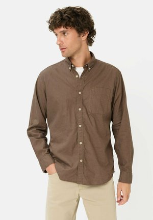 Man with curly hair wearing a brown button-up shirt over a white undershirt and beige pants, looking to the side with one hand in pocket.