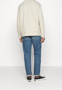 Personne portant une chemise à manches longues beige clair, un jean bleu retroussé et des baskets noires basses, debout devant un fond blanc uni.