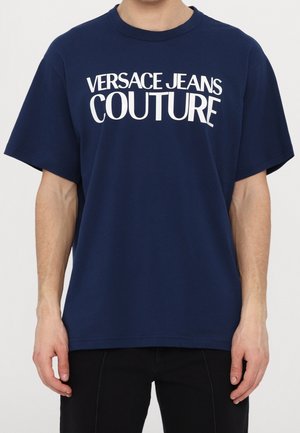 Man draagt een marineblauw T-shirt met witte "Versace Jeans Couture" tekst over de borst, gecombineerd met een zwarte broek.