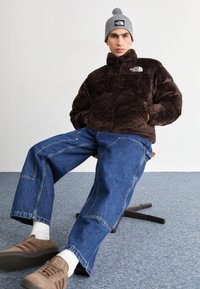 Brūns vilnas kapuce ar rāvējslēdzēju, zili plaša stila džinsi, pelēka adīta cepure ar pom-pomu un brūnas sporta kurpes ar teksturētām zolēm.