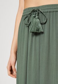 Jupe maxi verte et texturée avec une ceinture froncée, accentuée par un cordon de serrage avec des pompons. Vue rapprochée, mettant en valeur les détails et le tissu.