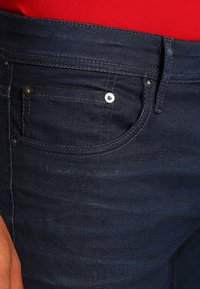 Dark denim jeans featuring a smooth texture, five-pocket design, and a small silver button detail on the front pocket.