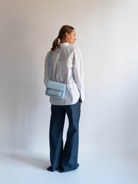 Lysblå rektangulær skuldertaske med klap og guldhardware, parret med en hvid skjorte og mørkeblå brede jeans.