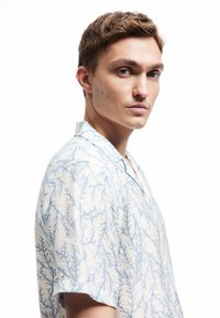 Chemise à manches courtes en tissu léger avec un fond blanc et un motif bleu ressemblant à des coraux ; possède un col et une coupe décontractée.