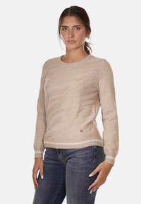 Suéter de punto beige con un sutil patrón de olas, cuello redondo, puños y dobladillo acanalados, y un pequeño logotipo metálico en el lateral.