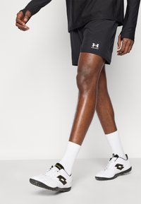 Μαύρο αθλητικό σορτς με λογότυπο Under Armour, συνδυασμένο με λευκές κάλτσες και λευκά αθλητικά παπούτσια με μαύρες και χρυσές λεπτομέρειες.