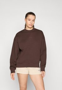 Sweatshirt marron oversize avec un col rond, présentant un texte brodé. Accompagné de shorts beige clair, texture lisse.