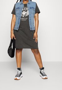 Denim bezrukávová bunda přes černé tričko s grafikou, spárovaná s tmavě šedými šaty a černými vysokými teniskami s bílými akcenty. Černá kabelka.