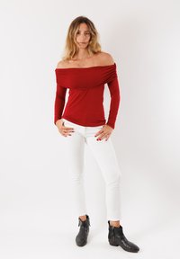 Top rosso scollato con spalle scoperte e maniche lunghe, dettaglio arricciato sul petto, abbinato a pantaloni bianchi slim-fit e stivaletti neri alla caviglia.