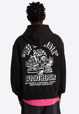 Person von hinten gesehen, trägt einen schwarzen Hoodie mit weißem Grafikdruck, der gestapelte Lautsprecher und den Text „Off the Wall Audio Repair“ sowie zusätzlichen kleineren Text zeigt.