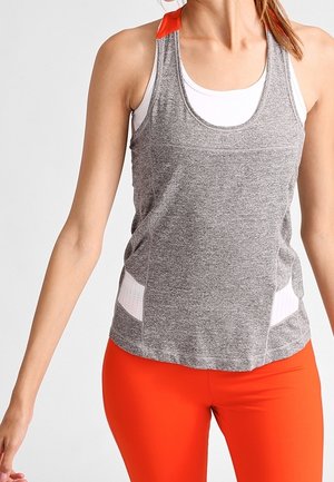 Kobieta ubrana w szary, bezrękawnikowy top sportowy na biały tank top i pomarańczowe legginsy, pozująca na jednolitym tle.