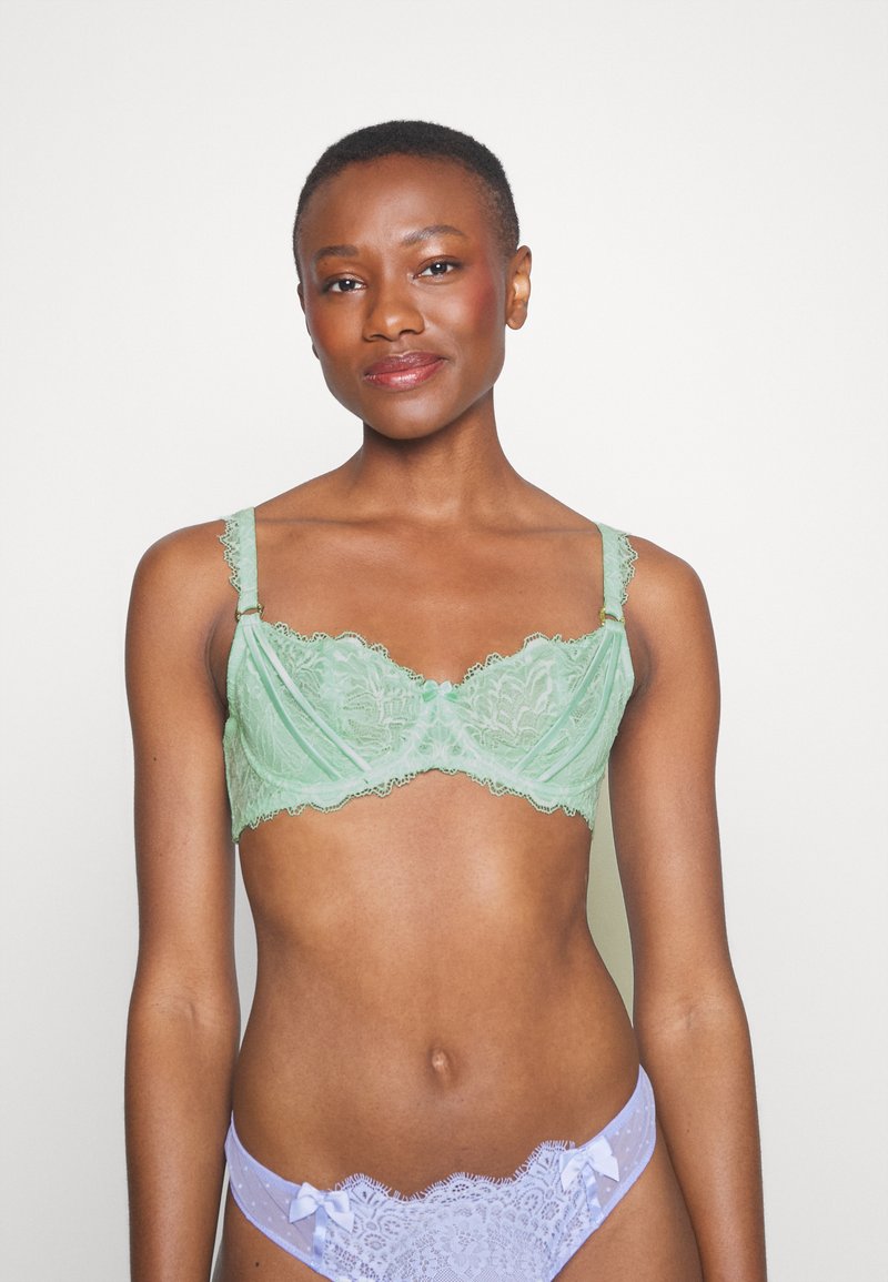 Zielony biustonosz koronkowy z falbaniastym wykończeniem, regulowanymi ramiączkami i dekoracyjną kokardką na środku. W połączeniu z lawendowymi koronkowymi majtkami.