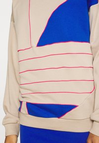 Svetr v béžové barvě s výraznými modrými a růžovými akcenty, který má geometrické vzory a volný střih. Strukturální látka s detailním prošíváním.