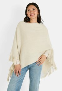Poncho de punto beige con bordes de flecos, que presenta un escote ancho y un acabado texturizado, llevado sobre unos jeans de color azul claro.