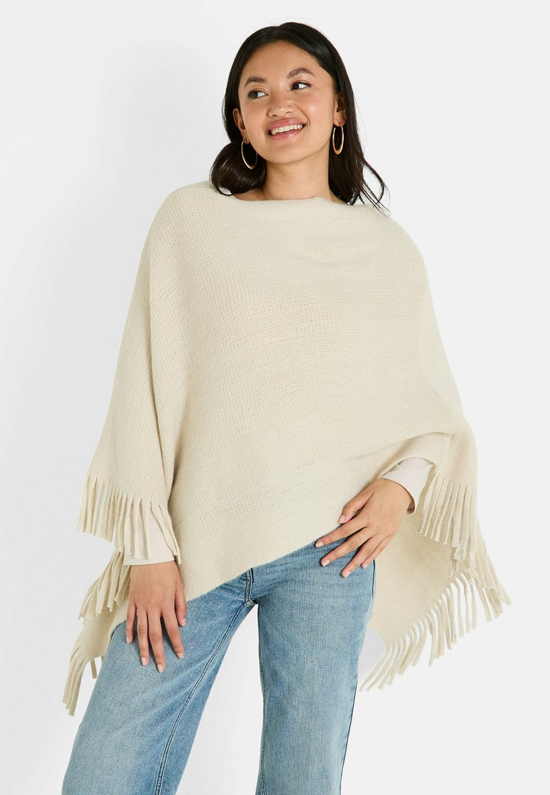 Poncho de punto beige con bordes de flecos, que presenta un escote ancho y un acabado texturizado, llevado sobre unos jeans de color azul claro.