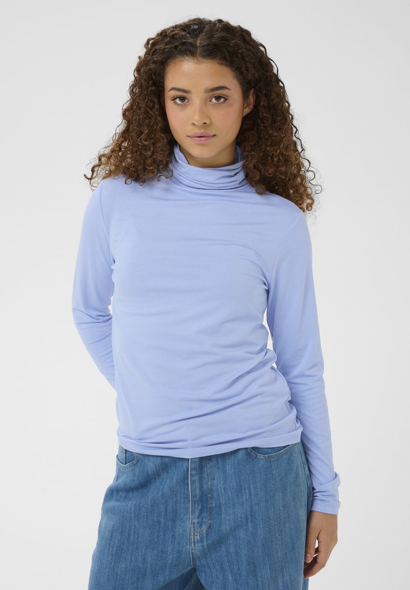 Jeune femme aux cheveux bouclés portant un col roulé bleu clair et un jean bleu, debout devant un fond clair uni.