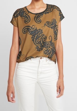 Vrouw draagt een bruine top met korte mouwen en een groot zwart-goud paisley-patroon, ingestopt in een witte broek met hoge taille.