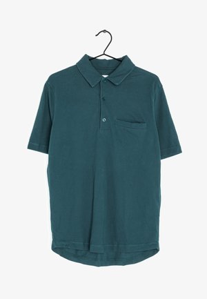 Camisa polo de manga corta en azul verdoso oscuro con collar, tres botones y un pequeño bolsillo en el pecho, colgando de una percha negra.
