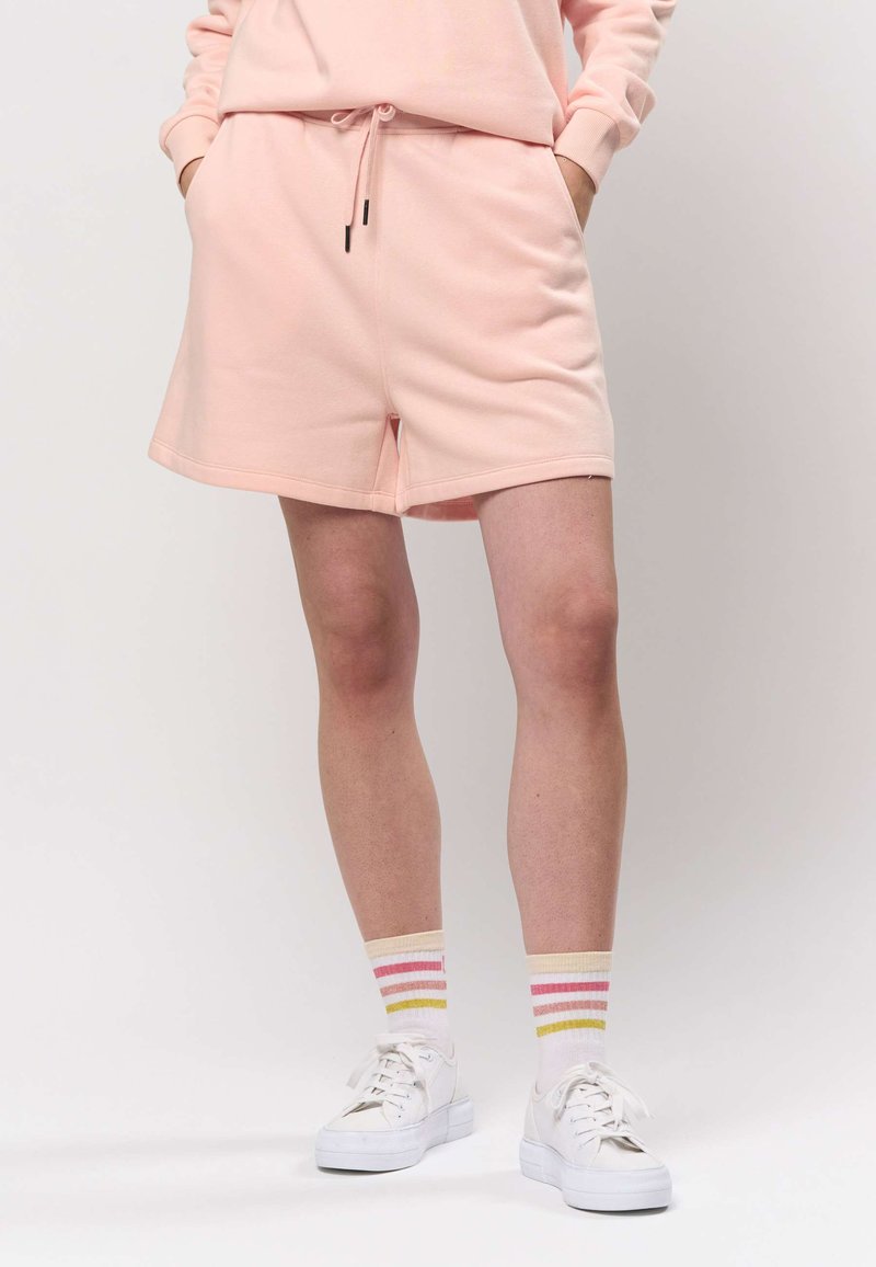 Světle růžové bavlněné šortky s elastickým pasem se stahovací šňůrkou, dvěma bočními kapsami a volným střihem. Kombinované s bílými teniskami a pruhovanými ponožkami.