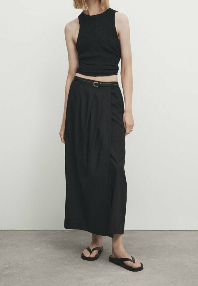 Femme portant un top court noir sans manches, un pantalon large noir à taille haute avec ceinture, et des sandales flip-flop noires, debout sur un sol gris.