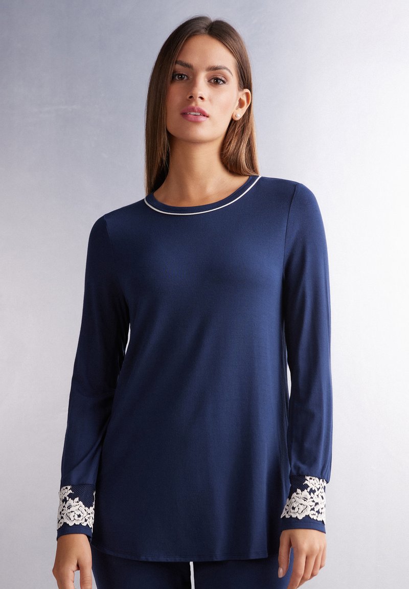 Marineblauwe langesleeve top van zacht materiaal met een ronde halslijn en crèmekleurige afwerking. De manchetten zijn versierd met witte bloemetjeskant.
