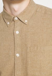 Hellbraunes Hemd mit Knopfleiste, strukturierter Stoff, spitzer Kragen, weiße Kunststoffknöpfe und eine Tasche auf der linken Brustseite.