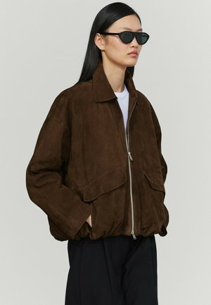 Femme aux longs cheveux noirs portant des lunettes de soleil foncées, une veste zippée en daim marron, une chemise blanche et un pantalon noir sur un fond uni.