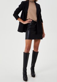 Blazer negro de doble botonadura sobre un cuello alto beige, combinado con una falda mini de cuero negra y botas de tacón alto negras hasta la rodilla.