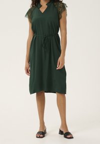 Zelené šaty délky ke kolenům s krátkými krajkovými rukávy a zavazováním v pase. Mají výstřih do véčka a boční rozparky pro snadnější pohyb.