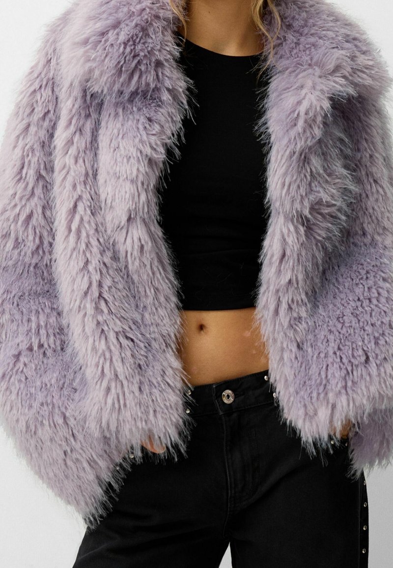 Jakke i imiteret pels i lys lavendel, med fluffy tekstur, bred krave og løs pasform, kombineret med en sort cropped top og sorte bukser.