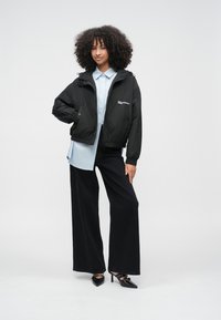 Mladá žena s kučeravými vlasmi, oblečená v čiernej bunde cez svetlomodrú košeľu na gombíky, široké čierne nohavice a čierne špicaté lodičky, stojaca sebavedomo.