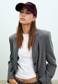 Bordeauxrotes Samt-Baskenmütze mit gesticktem Text, kombiniert mit einem eng anliegenden weißen T-Shirt und einem grauen Nadelstreifenblazer. Das Model hat lange Haare.