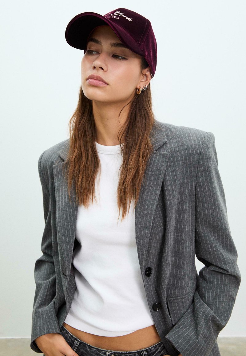 Bordeauxrotes Samt-Baskenmütze mit gesticktem Text, kombiniert mit einem eng anliegenden weißen T-Shirt und einem grauen Nadelstreifenblazer. Das Model hat lange Haare.